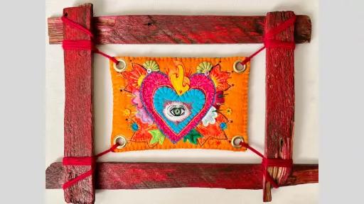 A rustic red wooden frame with a piece of felt art suspended in the middle of the frame showing a heart with an eye in the centre and flame coming from the top.