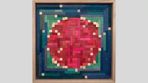 A framed piece of art made of wooden rectanges arranged to make a mainly pink circle surrounded by a square of greens and blues.