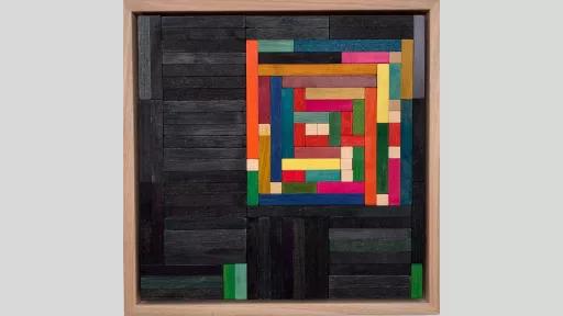 A framed piece of art made of wooden rectanges arranged to make a suare of rainbow blocks, surrounded by dark grey blocks with some small green blocks on the edge of the piece