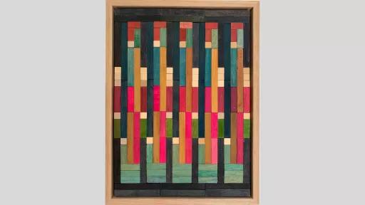 A framed piece of art made of wooden rectanges arranged in columns with bright pink, blue and some light and dark brown pieces in the middle of the columns surrounded by dark grey pieces