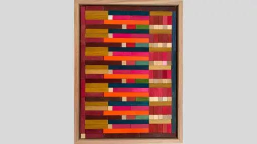 A framed piece of art made of wooden rectanges arranged in rows horizontally across the piece with bright ornages and pinks used alongside a few brown, green and blue pieces