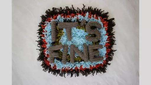 A piece of carpet with black, red, light blue, and green wool in concentric circles, overlaid with crocheted letters spelling out 'It's fine'