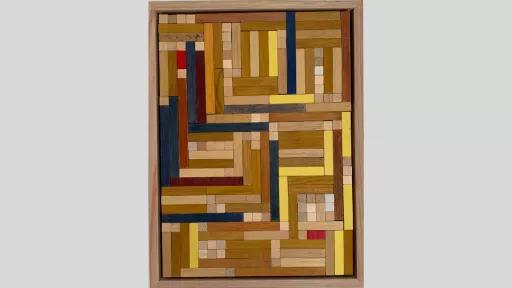 A framed piece of art made of wooden rectanges arranged to tesselate with each other changign from horizontal to vertical throughout, and using mostly light brown pieces along with some yellow and blue and a few red