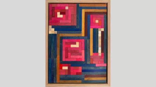 A framed piece of art made of wooden rectanges arranged pink squares of varying sizes with mostly blue blocks between the squares