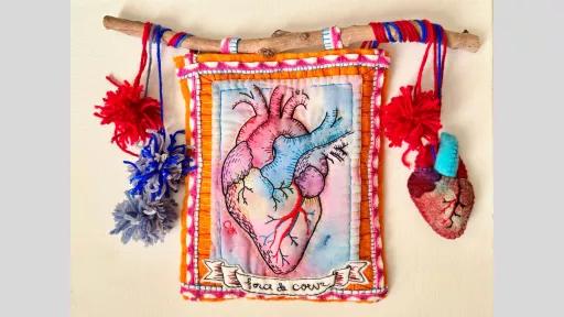 A piece of artwork on material of a anatomically correct heart embroidered with rainbow accents adn with the text 'force de coeur' in a banner along the base. The artwork is hanging from a stick with wool, and there are pompoms and a felt heart hanging from the wool.