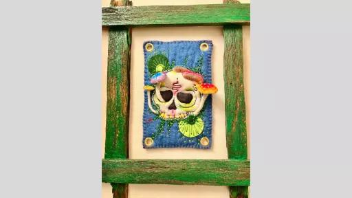 A rustic green wooden frame with a piece of felt art in the middle of the frame showing a skull with mushrooms growing out of the skull which are raised from the felt so they emerge from the piece.