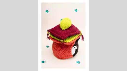 A small stack of crocheted pillows on a small kettle with a crocheted cover on it, sitting on a background with little blue flowers