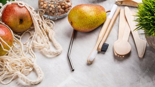 A close-up of a table with pasta, fruit, and nuts all in sustainable containers or bags, and wooden cutlery and toothbrushes with a metal straw and a plant alongside