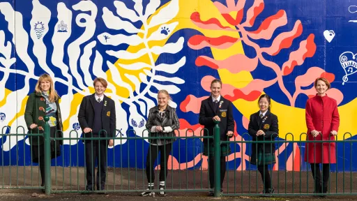 A group of 3 students and 3 adults standing in front of a bright blue mural with mutlicoloured patterns of plants