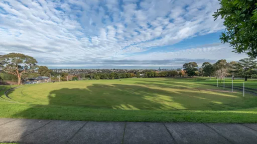 Oval-shaped grass field surrounded by two large grass and brick steps. There are trees and houses are in the distance and a footpath in the foreground.