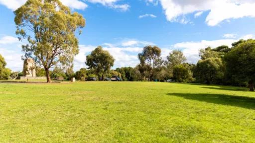 Grass area with parked cars and trees in the distance. There is a partly obscured concrete memorial structure to the left.