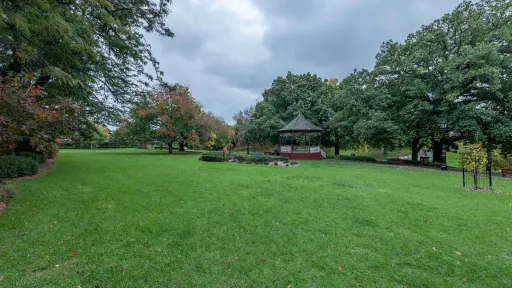 Large slightly sloped grass area with a small garden in the middle and trees around the edge. There is a rotunda and footpath on the far edge.