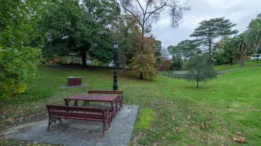 Table with two bench seats next to a light post and barbecue, in a grass area with mature trees. There is a small valley and walking bridge to the right.