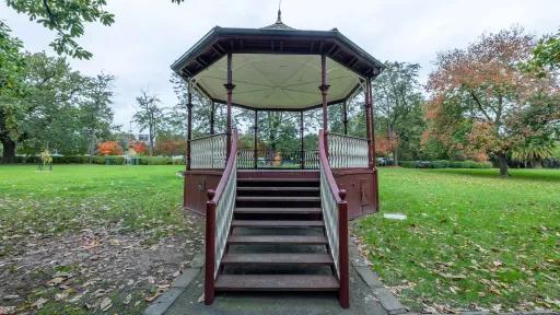 Steps up to dark red rotunda with white balustrades and roof, located in a grass area. There are mature trees are in the distance.