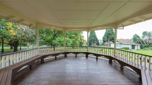 Inside view of round outdoor rotunda in grass area, with trees nearby and a white house to the right. The column, roof and balustrades are white, and the floor and bench seats are dark red.