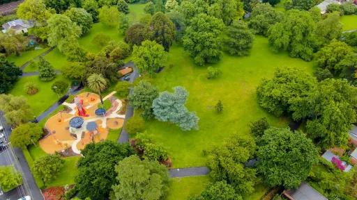 Aerial view of grass area with many scattered green trees. There is a large playground with surrounding footpaths to the left.