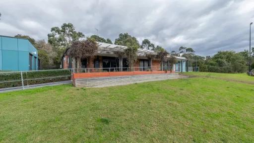 Single-storey brown brick building on the edge of a grass field. Thick shrub is growing in several places along the roof. There is part of a green building to the right and trees in the distance.