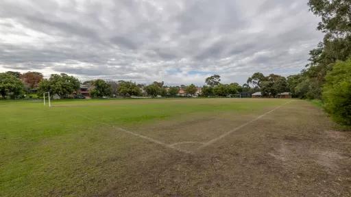 Corner view of soccer pitch under grey sky with goals at each end and medium trees on the horizon. There is a large bare dirt patch in the foreground.