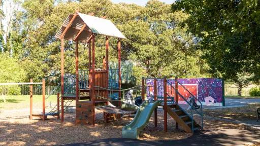 Playground featuring wooden climbing structure with triangular corrugated roof. There is a purple, brown and white mural in the background and trees in the distance.