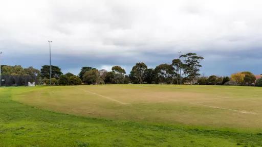 Corner view of soccer pitch with white markings. There is thicker grass in the foreground and tall trees on the horizon. Cricket nets are visible to the far left.