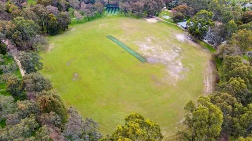 Aerial view of oval field with cricket pitch and some bare patches, surrounded by a thick layer of trees. There are cricket nets at the top and a small building to the top right.