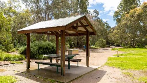 Table and bench seats under a small wooden structure with a triangular tin roof. There is a barbecue alongside the grass and trees in the distance.