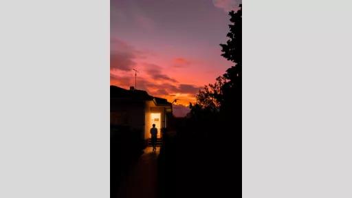 Silhouette of house at dusk with person standing in front of an illuminated front door. There is a tree to the right.