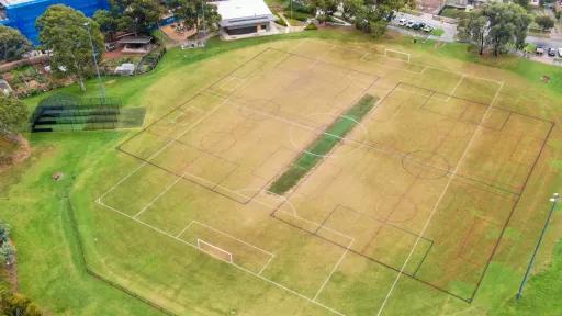 Aerial view of sports field with white markings for soccer and black markings for another sport. There are cricket practice nets to the left, a pavilion at the top and a road to the top right.