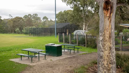 Barbecue and two sets of tables and chairs between a grass field and a dirt path. There is a tree trunk to the near right, a fenced-off nursery in the background and cricket nets in the distance.