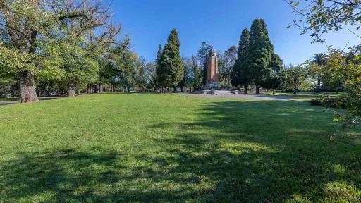 Area of lush grass in sunshine with concrete memorial structure and tall trees at the far end. There are shadows in the foreground.