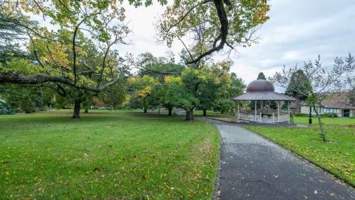 Large grass area with a rotunda and walking path to the right. There are scattered medium-sized trees and fallen leaves on the ground.