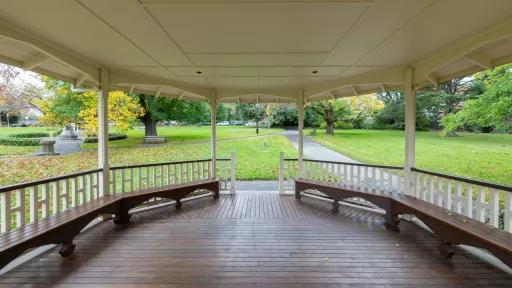 Inside view of rotunda in grass area. The columns and bench seats are dark red and the roof and balustrades are white. There are paths, a park bench and a round hedge garden in the background.