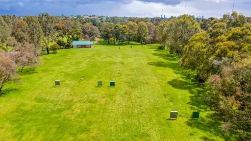 Large rectangular grass area surrounded by a thick layer of trees. A small single-storey building is at the far end. There are five archery targets in the foreground.