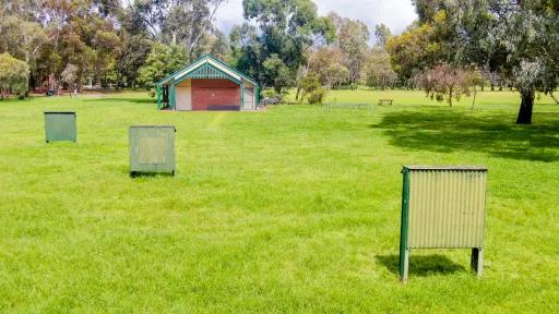 Three archery targets in staggered locations in green grass. There is a small building at the far end and trees in the background.