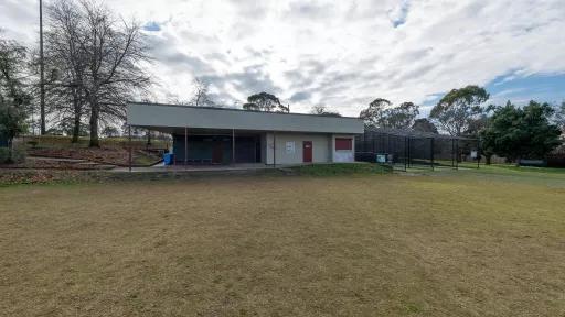 Single-storey light and dark grey building on the edge of a sportsground. There are cricket nets to the far right and a tall bare tree and light post to the left.