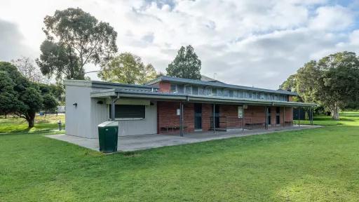 Single-storey brown brick building with covered concrete patio and rubbish bin, on the edge of a grass field. There is an adjoining corrugated structure with roller door to the left.