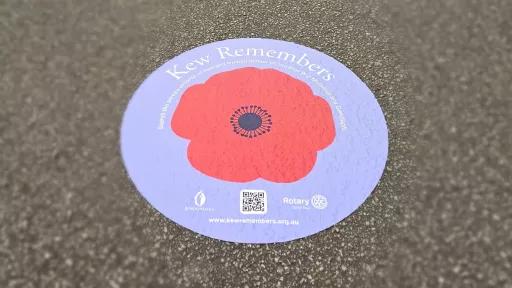 A floor decal on pavement, displaying a red poppy on a purple background with the heading 'Kew Remembers' and a qr code