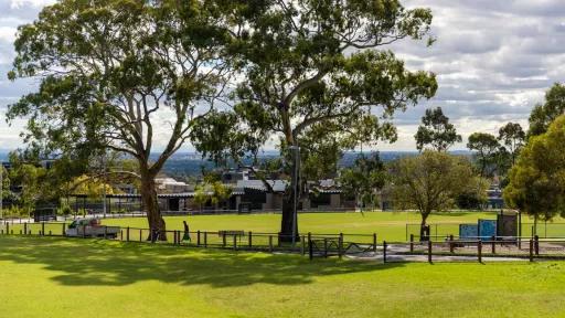 A sports oval with green grass. The oval is surrounded by a wire fence and tall trees surround the oval. A building and an undercover area is right next to the oval.