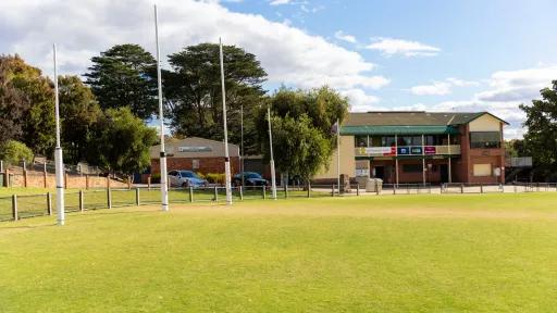 Part of an oval grass field with tall trees and white goalposts to the right. There are two house-sized buildings on the field edge.