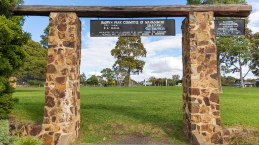 Rectangular entrance structure for grass field, with two brick columns, two information signs and a wooden plank. There are tall trees are in the distance.