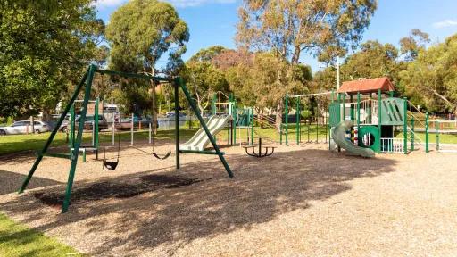 Playground on a mulch surface. It has swings and two climbing features with slides, surrounded by a grass area and tall trees.