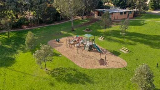 An overhead image of a playground with a mulch floor. It is surrounded by a large grass area. A walking path runs along the edge separating the park from a tree area. A brick building is also in the background. 