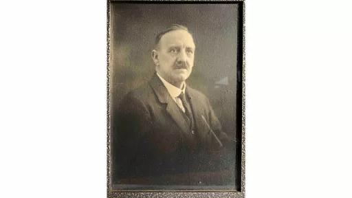 Framed photograph of a man with combed back hair and moustache, wearing a black suit, vest, and tie with a white collared shirt. The man has been photographed from the waist up and is looking straight ahead, eyes looking past the camera lens. 