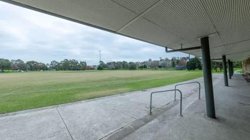 Looking out towards a green sports field from a sports grandstand . Trees and houses are visible in the background. 