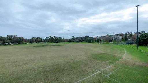 A wide, grassy field. The field is surrounded by trees and houses and there are light towers at either end of the field.