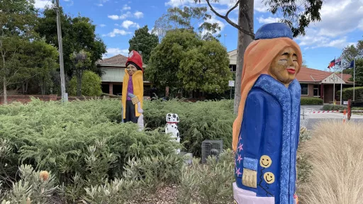 A garden bed with low shrubs and 3 wooded carved sculptures. 1 of a dalmation, and 2 people, one a fireman and the other a woman wearing a long blue coat and matching hat.