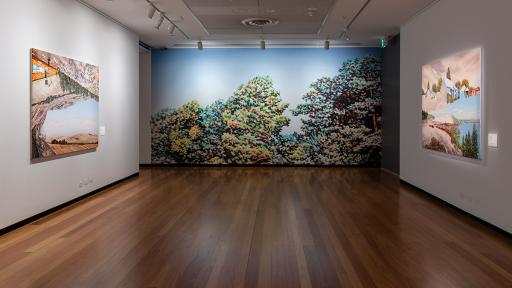Photograph of large landscape paintings hanging in a gallery. At the end of the gallery is a large print of painted tree tops
