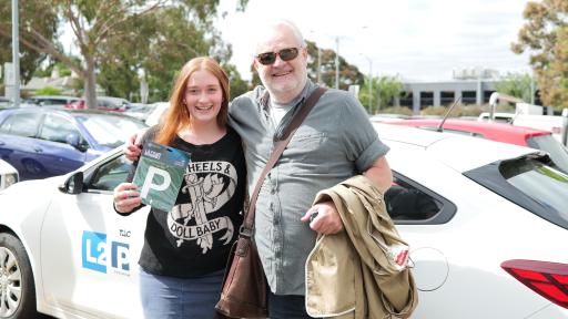 Young woman holding a P plate next to older man and in front of a car