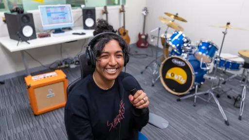 A smiling woman holds a microphone with a drumkit behind her