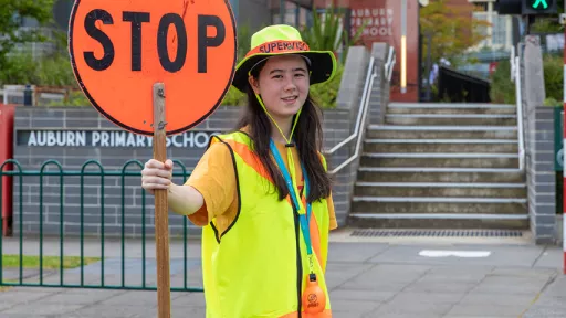 School crossing supervisor wearing hi-vis hat and vest holds stop sign in front of steps leading to Auburn Primary School.